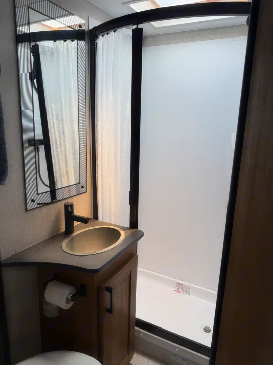 Inside an RV rental, a functional bathroom features a sleek sink, black faucet, mirror, and curved shower with translucent curtain.. Coachman Leprechaun 2025