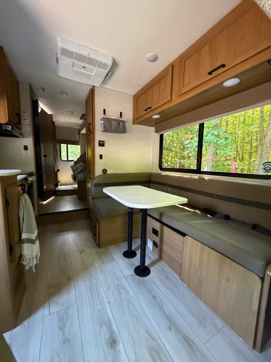 RV rental interior features a bright dinette area, kitchen, and a window framing a lush forest view, offering a cozy camping experience.. Coachman Leprechaun 2025