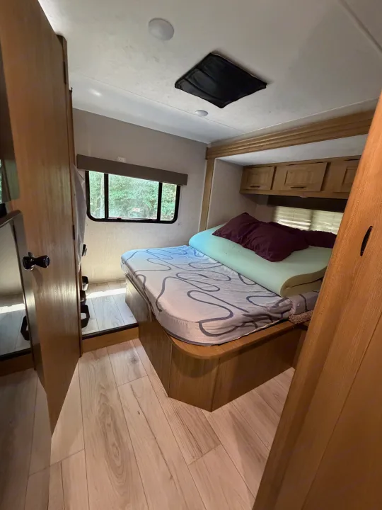 Compact rv rental bedroom: bed with memory foam, wood interior, cabinets, and window view of trees.. Coachman Leprechaun 2025