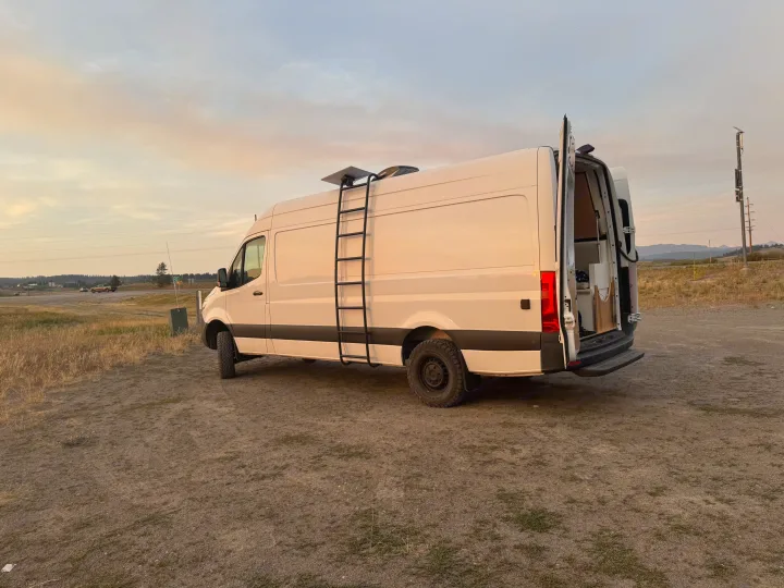 White rv rental camper van, doors open, on dirt. Features a ladder and roof tech for off-grid travel & van life adventures.. Mercedes Sprinter 2022