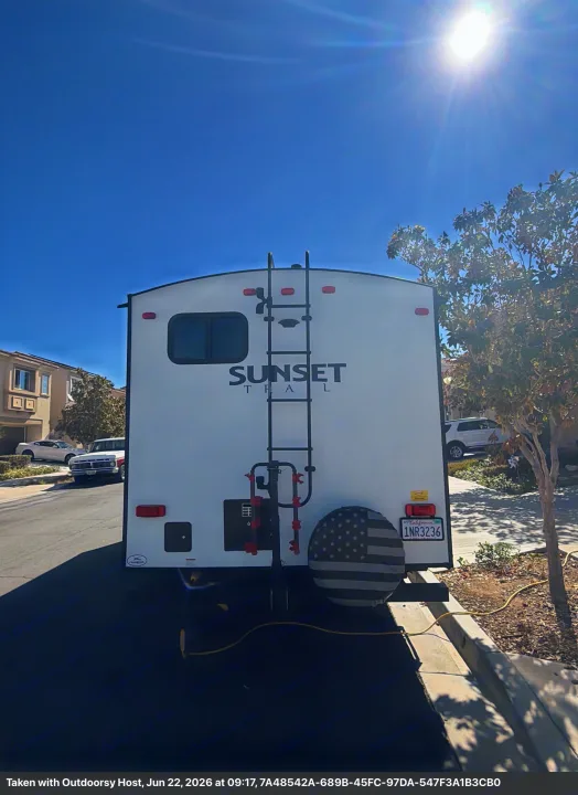 Rear of a Sunset Trail rv rental, parked on a sunny residential street. Features a ladder and USA-themed spare tire cover.. Crossroads RV Sunset Trail Super Lite 2021