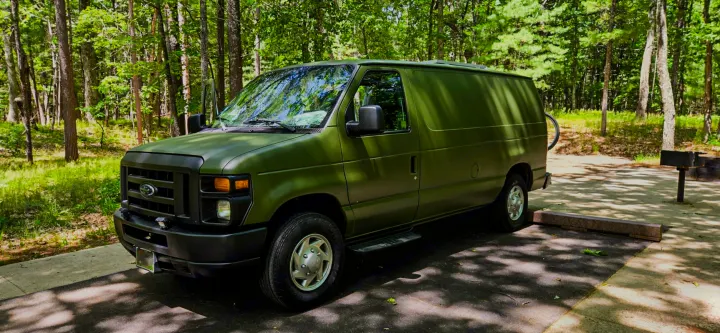 Olive green Ford E-Series van, likely an RV rental, parked at a wooded campsite with dappled sunlight.. Ford Econoline 2012