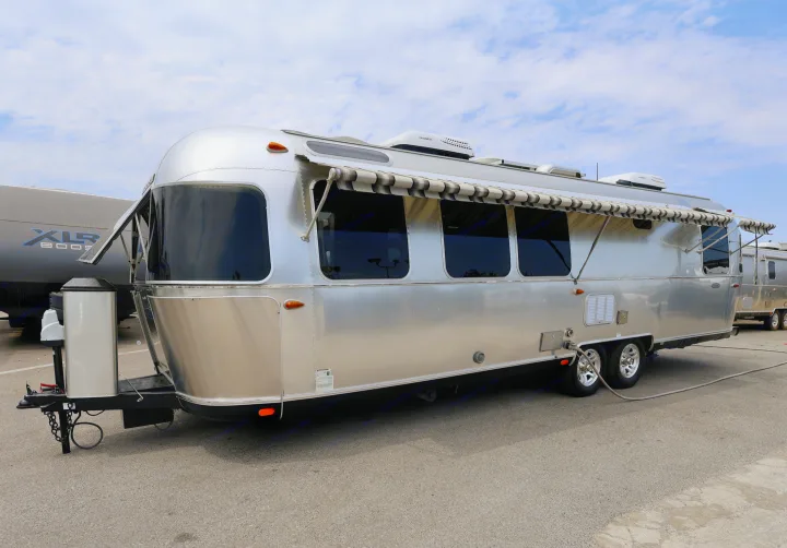 This silver Airstream travel trailer, ideal for an RV rental, features dark windows and an extended awning, parked under a cloudy sky.. Airstream Classic 2019