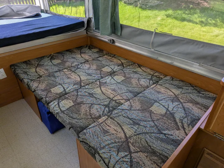 Interior view of an RV rental showing a blue mattress on a bed, colorful patterned dinette cushions, and a screened window.. Coleman Destiny Sedona 2002