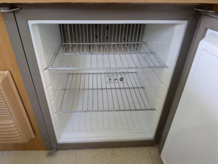 Interior of an empty RV rental refrigerator, featuring wire shelves and exposed metal cooling fins at the back.. Coleman Destiny Sedona 2002