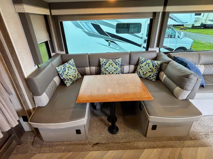 Comfortable RV rental dinette with grey U-shaped seating, patterned pillows, and a wooden table by large windows.. Forest River Wildwood 2021
