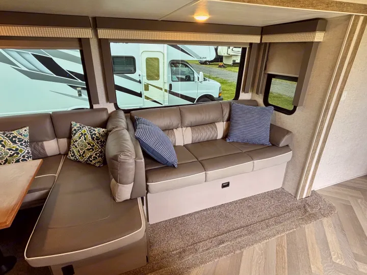 An inviting RV rental interior showing a grey L-shaped sofa with pillows, a partial table, and bright windows looking outside.. Forest River Wildwood 2021