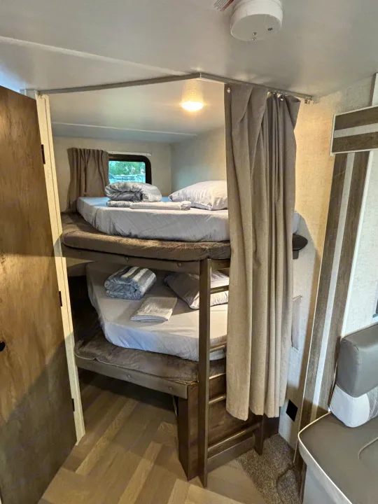 Cozy bunk beds in an rv rental, featuring light gray bedding, privacy curtains, and a window for comfortable sleeping space.. Forest River Wildwood 2021