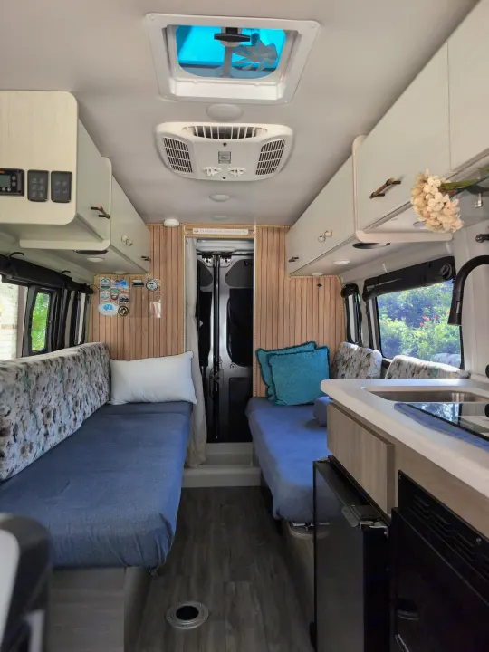 Bright rv rental interior: two blue-covered twin beds, kitchen counter, overhead cabinets, wooden walls, and ceiling fan/AC. Cozy camper setup.. Thor Tellaro 2024