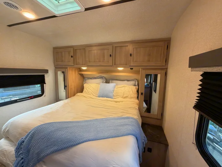 Cozy RV rental bedroom with a comfortable queen bed, light wood cabinets, mirrors, and windows with blinds.. Coachmen 2963BH 2023