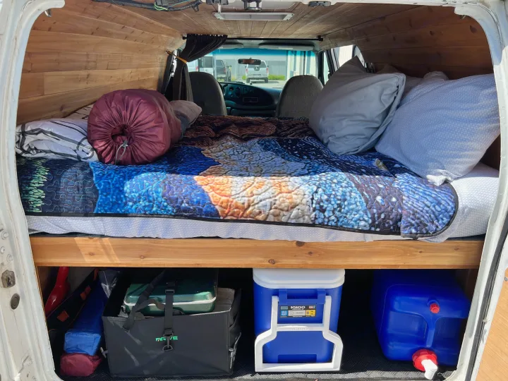 RV rental van interior. A cozy bed with colorful quilt, pillows, and under-bed storage (cooler, water jug).. Ford Econoline 150 1997
