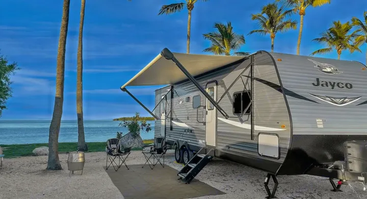 Coastal rv rental setup: a Jayco travel trailer with an awning and chairs by the ocean, flanked by palm trees.. Jayco Jay Flight SLX 2021