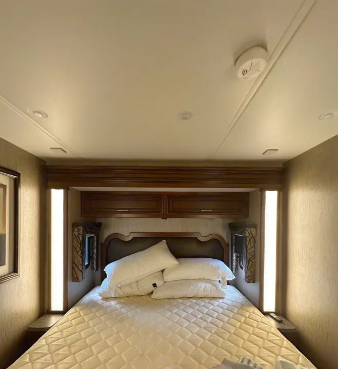 Inside this RV rental bedroom, a quilted bed with stacked pillows, dark wood cabinets, and integrated lighting create a cozy space.. Newmar Canyon Star 2016