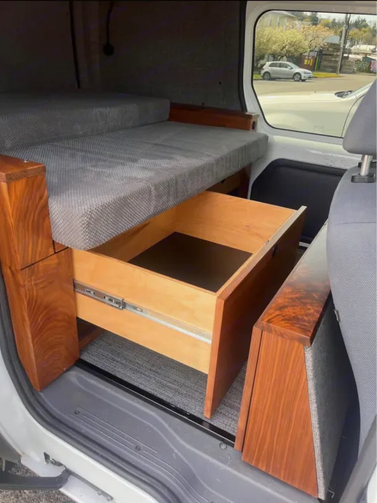 Inside an rv rental: a grey bed, dark wood accents, and a pulled-out storage drawer with light wood interior, visible from the door.. Ford Transit 2013