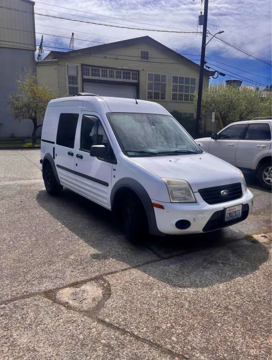 A white Ford Transit Connect van, ideal for an rv rental, parked on concrete. Industrial building & cranes are in the background.. Ford Transit 2013