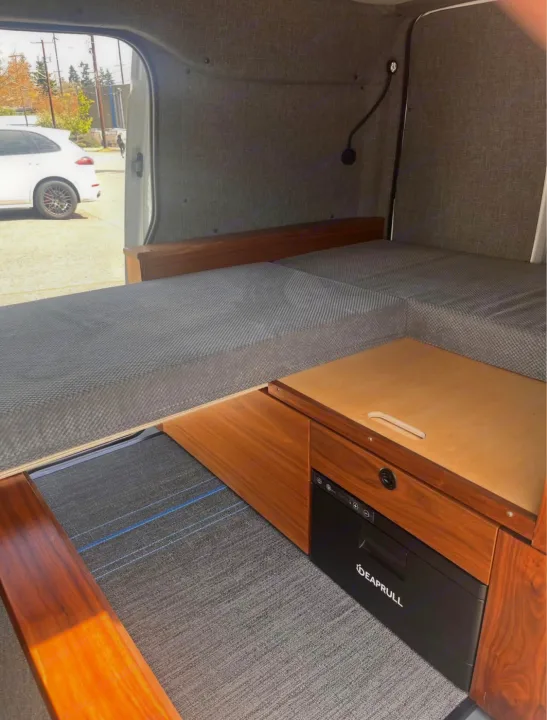 Inside a custom rv rental: cozy camper van interior with a grey bed, wood cabinetry, built-in fridge, and reading lamp.. Ford Transit 2013
