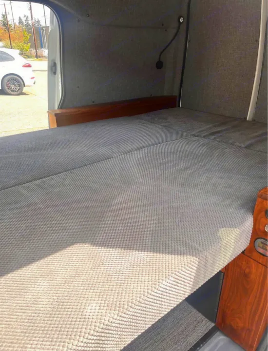 RV rental interior: A comfortable grey textured bed and wooden accents inside a camper van, with the door open to the outdoors.. Ford Transit 2013