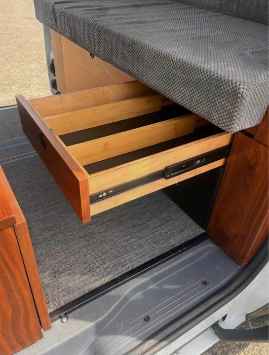 Explore this RV rental's custom camper van storage: a pull-out wooden drawer with dividers under a textured gray bed.. Ford Transit 2013