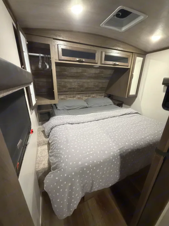 Cozy RV rental bedroom featuring a bed with grey patterned bedding, wood paneling, overhead cabinets, and a ceiling vent.. Keystone RV Cougar 2023