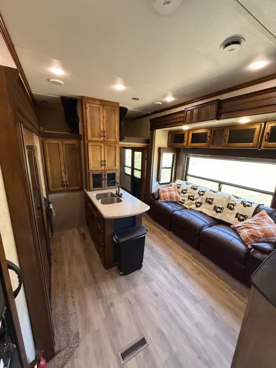 Spacious RV rental interior featuring a modern kitchen island with wood cabinetry, a comfortable brown leather sofa, and light wood-look flooring.. Keystone RV Carbon Toy Hauler 2015