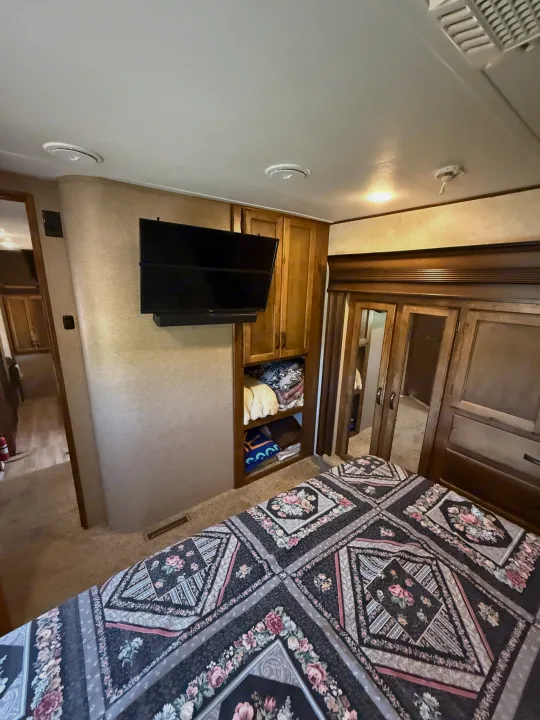 RV rental bedroom with a detailed floral bedspread, wall-mounted TV, and built-in wooden storage cabinets.. Keystone RV Carbon Toy Hauler 2015