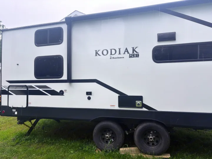 White Kodiak SE travel trailer (RV rental) side view, parked on grass in light rain. Features dual axles and black trim.. Dutchmen Kodiak SE 2021