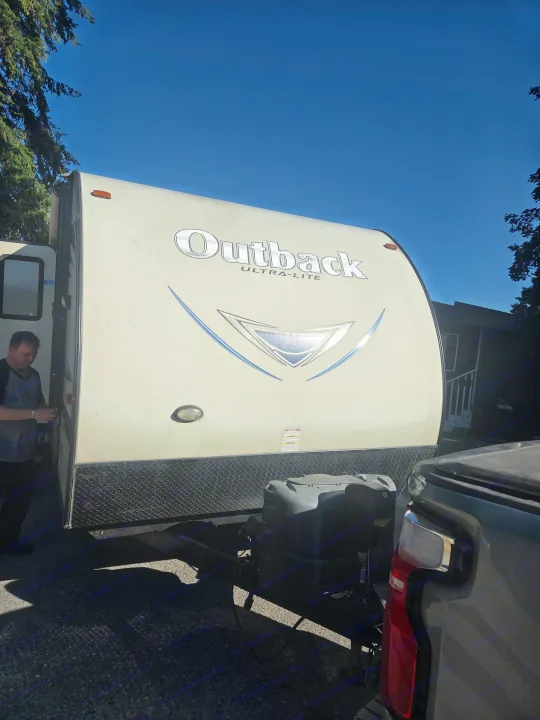 An RV rental, an Outback Ultra-Lite travel trailer, is hitched to a truck. A man stands by the RV's door under a blue sky.. Keystone Outback 2017