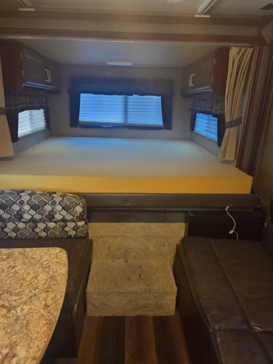 RV rental interior: elevated sleeping area with foam mattress & overhead cabinets, windows, and steps leading to dinette seating.. Keystone Outback 2017