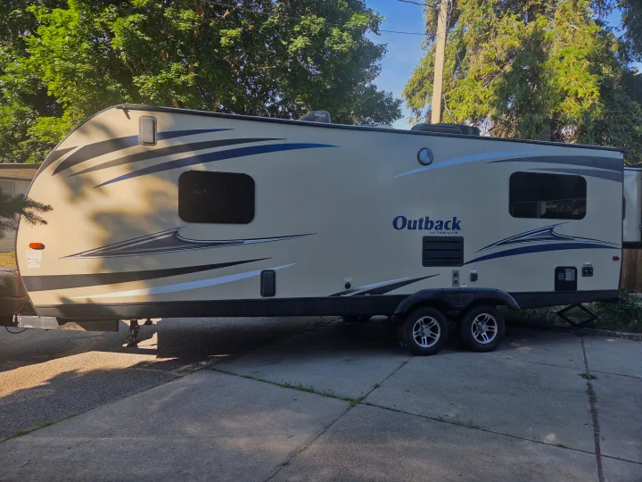 Outback Ultra-Lite travel trailer, perfect for an RV rental. Parked on a concrete driveway with trees in the background.. Keystone Outback 2017