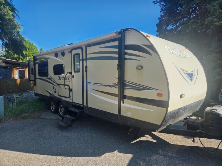 Outback Ultra-Lite RV rental: A tan travel trailer with black/white graphics, parked on asphalt at a sunny campsite.. Keystone Outback 2017