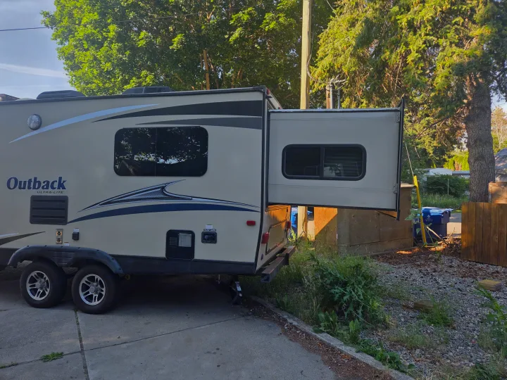 Beige Outback Ultra-Lite RV rental with slide-out extended, parked on a driveway near trees and utility bins.. Keystone Outback 2017