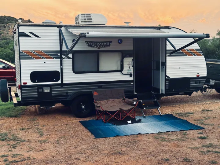 Relaxing campsite scene: a white travel trailer RV rental with an extended awning, a chair, and blue mat on gravel at sunset.. Forest River Wildwood X-Lite 2021