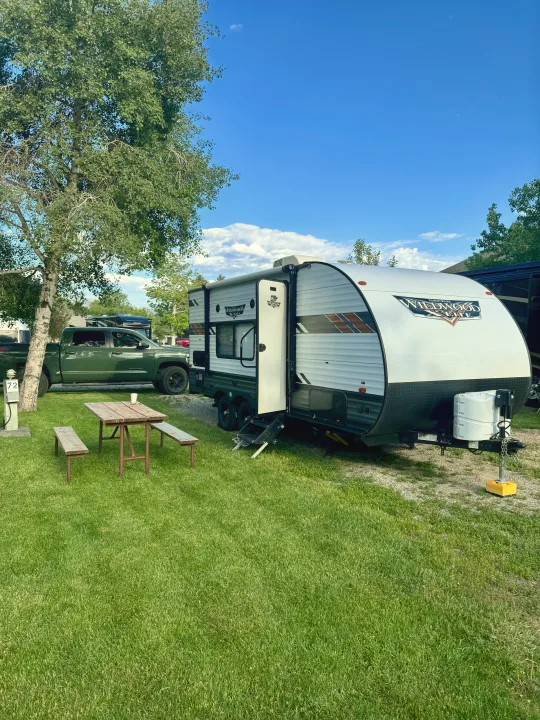 This RV rental, a white travel trailer, is parked at a sunny campground with a green pickup truck, picnic table, and lush grass.. Forest River Wildwood X-Lite 2021