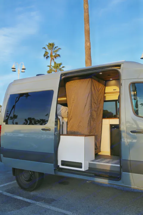 Open side door of an RV rental revealing its built-out camper van interior with a bed area, kitchen, and curtain.. Mercedes-Benz Sprinter 2023
