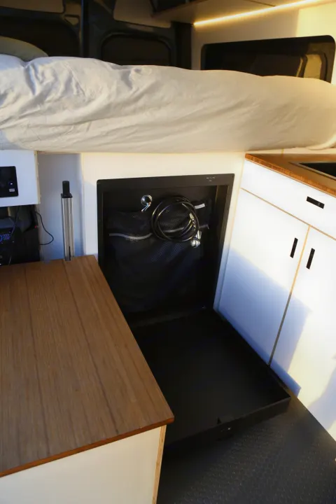 This RV rental interior features a cozy bed, wooden counter, and smart pull-out storage with cables for efficient space.. Mercedes-Benz Sprinter 2023