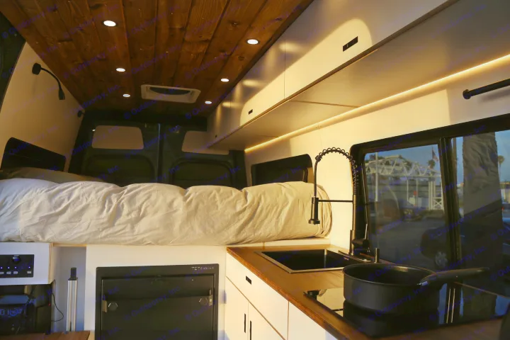 Cozy RV rental interior: A raised bed, modern kitchen with sink/stove, and warm ambient lighting for comfortable van life.. Mercedes-Benz Sprinter 2023
