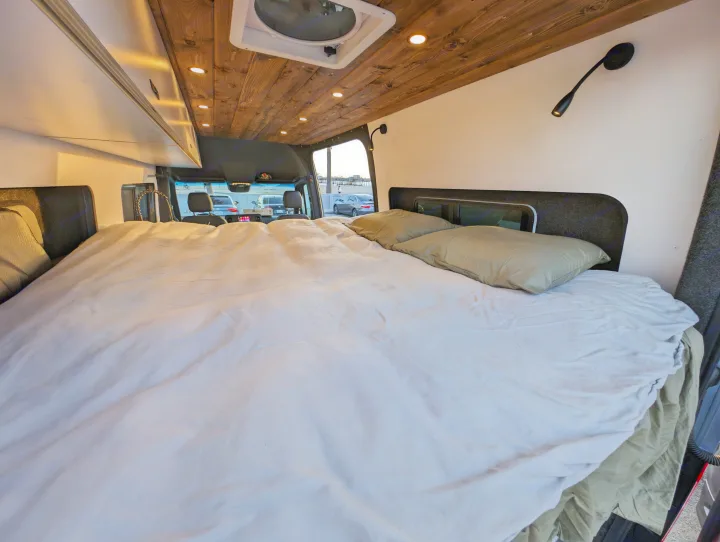 RV rental interior: comfy bed with light bedding, wooden ceiling with lights, and a scenic view of cars and a distant pier.. Mercedes-Benz Sprinter 2023