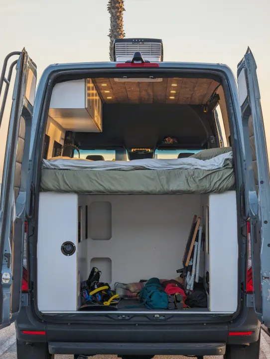 The rear of a custom rv rental camper van, doors open, revealing a cozy bed above a storage area filled with adventure gear.. Mercedes-Benz Sprinter 2023