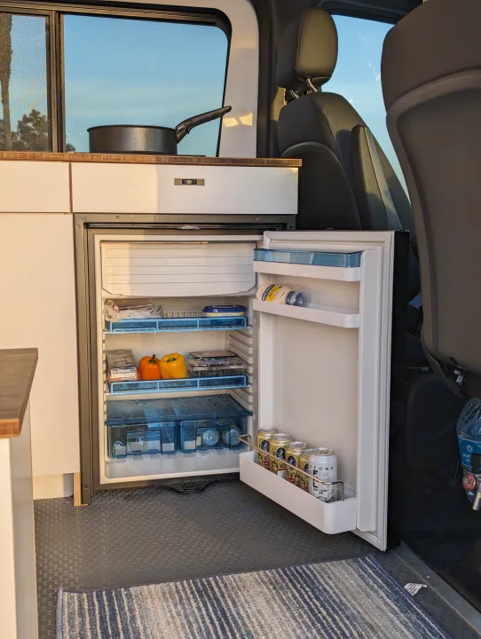 RV rental interior: Open fridge with food & drinks in a camper van kitchen. Ideal storage for travel adventures!. Mercedes-Benz Sprinter 2023