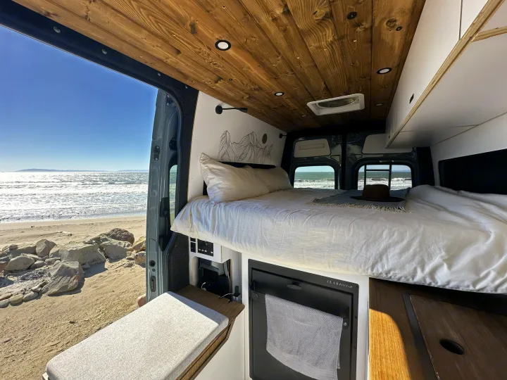 Cozy RV rental camper van interior, featuring a comfortable bed, fridge, and incredible ocean views from a sandy beach.. Mercedes-Benz Sprinter 2023