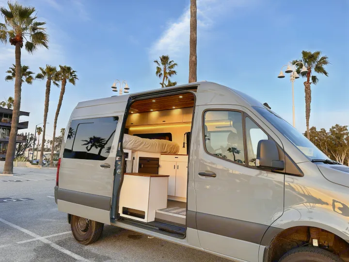 Modern grey camper van with its door open, showcasing a cozy, custom interior. Parked with palm trees in the background, ideal for an rv rental.. Mercedes-Benz Sprinter 2023