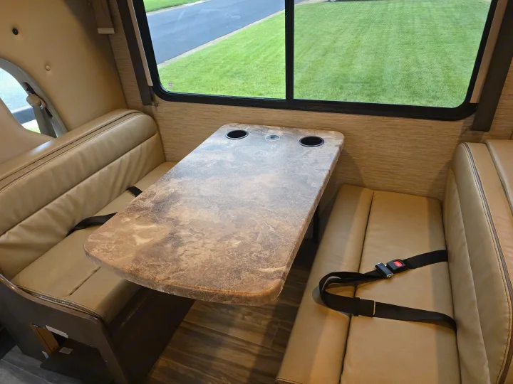 Inside an rv rental: a cozy dinette with a table, cup holders, and safety seat belts. Green view out the window.. Thor Four Winds 2023