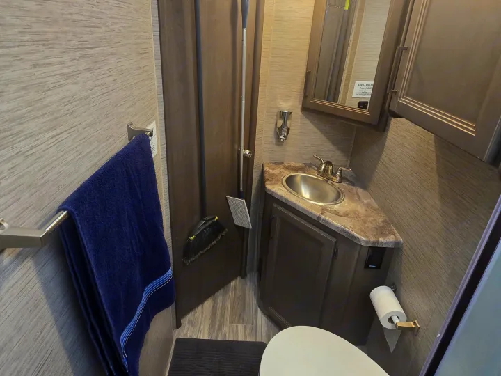 Inside an RV rental bathroom, showing a compact vanity with sink, toilet, mirrored cabinet, blue towel, broom, and dustpan.. Thor Four Winds 2023