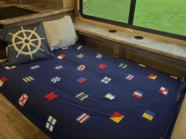 Cozy RV rental interior features nautical flag bedding, a ship's wheel pillow, and window with built-in cupholders.. Thor Four Winds 2023