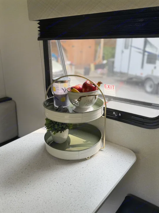 Bright RV rental interior. A tiered stand holds fresh apples, a lavender candle, and a small plant on a speckled countertop by the window.. Heartland's RV Mallard 17 BHL 2025