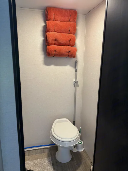 Compact rv rental bathroom with white toilet, stacked orange towels, and cleaning supplies. Efficiently designed for travel.. Heartland's RV Mallard 17 BHL 2025