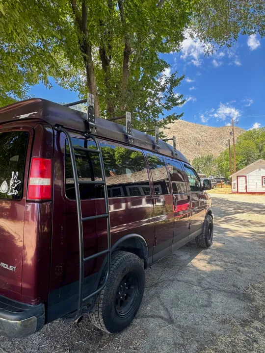This dark red Chevy camper van, equipped with a ladder & roof rack, could be an RV rental, ready for mountain adventures.. Chevrolet Open Road 2005