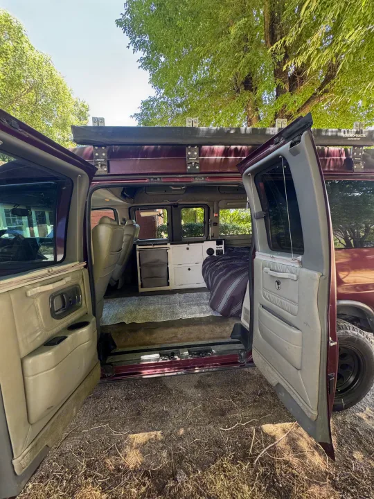 Converted camper van interior with doors open, showing bed & storage. Ideal for an RV rental outdoor adventure under green trees.. Chevrolet Open Road 2005