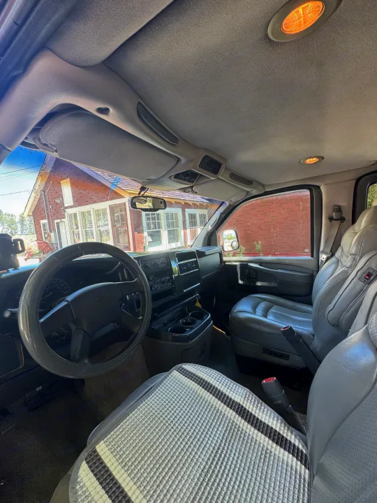 RV rental interior: Driver's cabin view featuring steering wheel, dashboard, and grey leather seats. A brick house is visible outside.. Chevrolet Open Road 2005