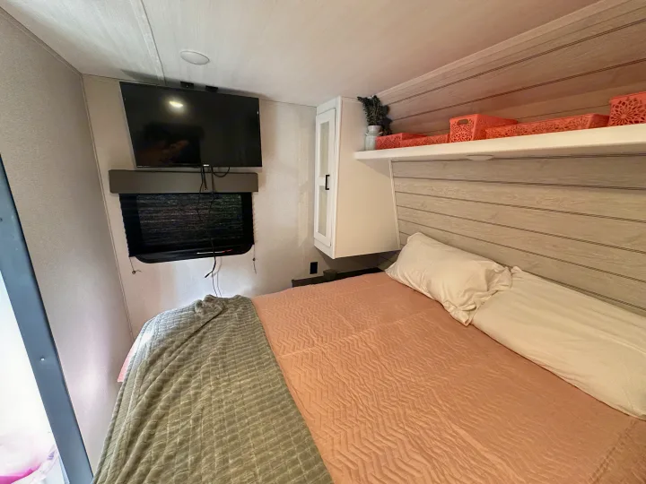 This RV rental's bedroom offers a cozy bed, wall-mounted TV, and functional overhead storage with pink accents.. Keystone RV Hideout 2023